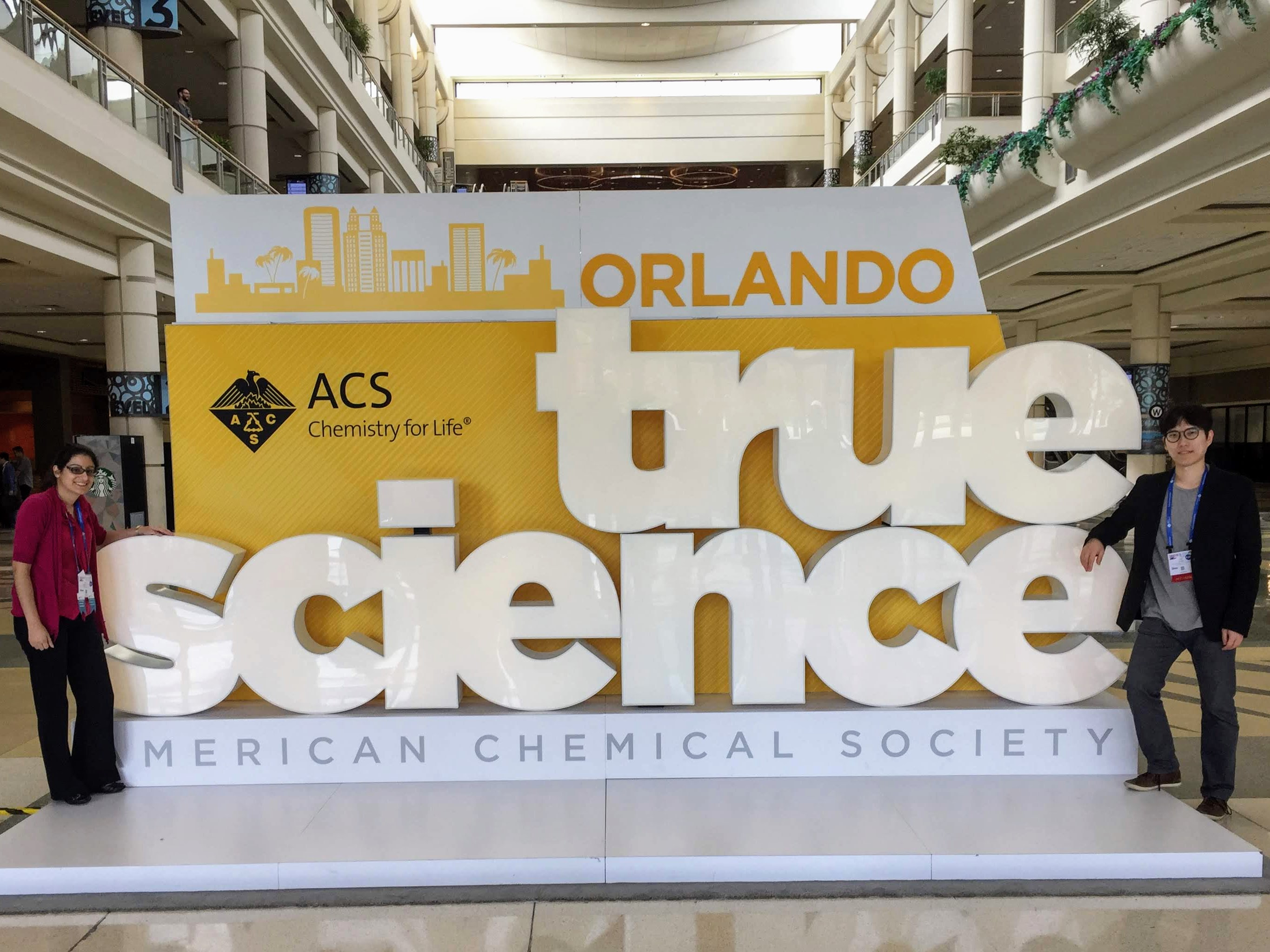 One female student standing to the left and one male student standing to the right of a large white lettered sign that read 'true science American Chemical Society'